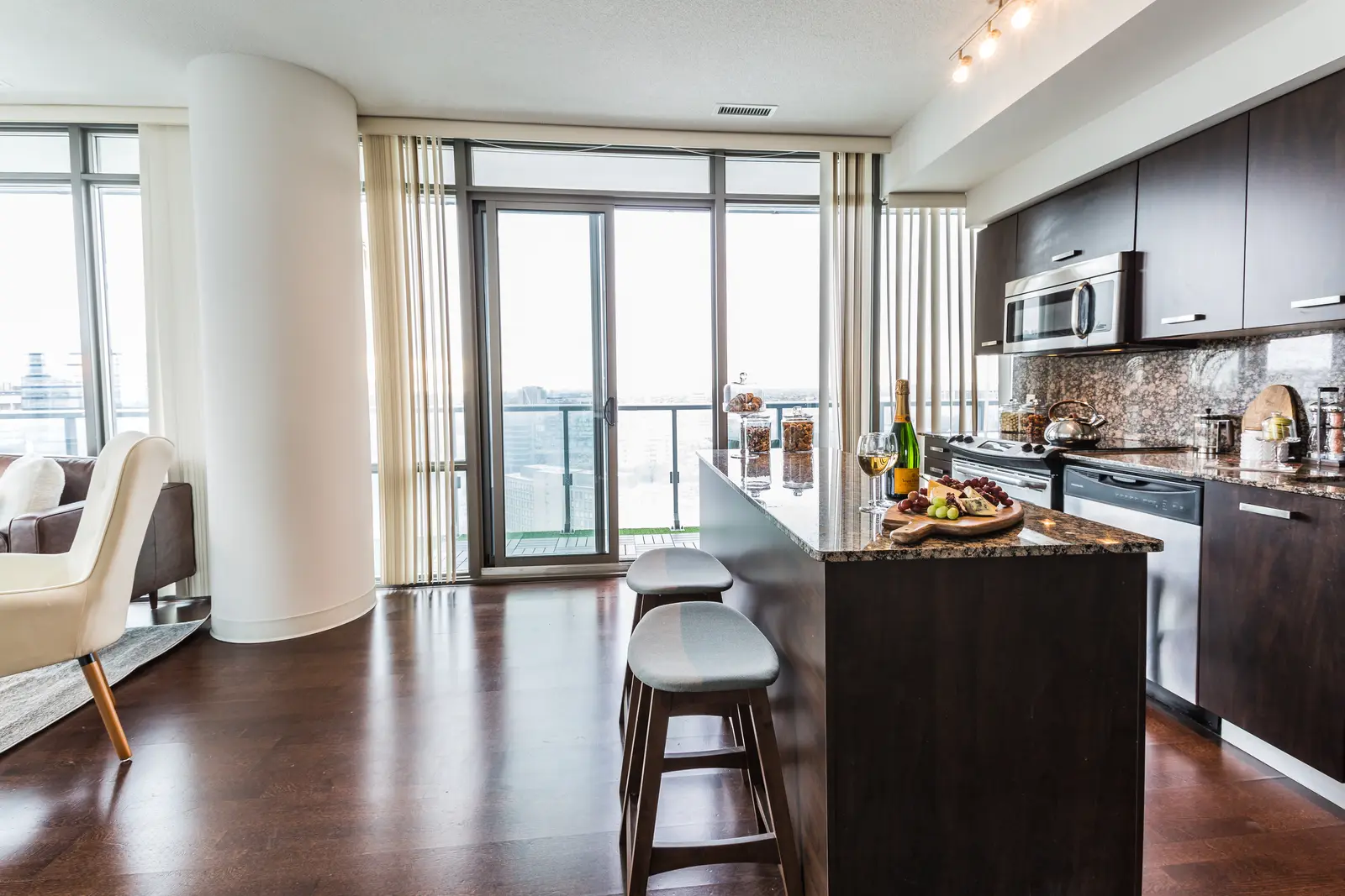Interior of a Bay Street condo in Toronto, with a grey sofa, a dark kitchen, and city windows.