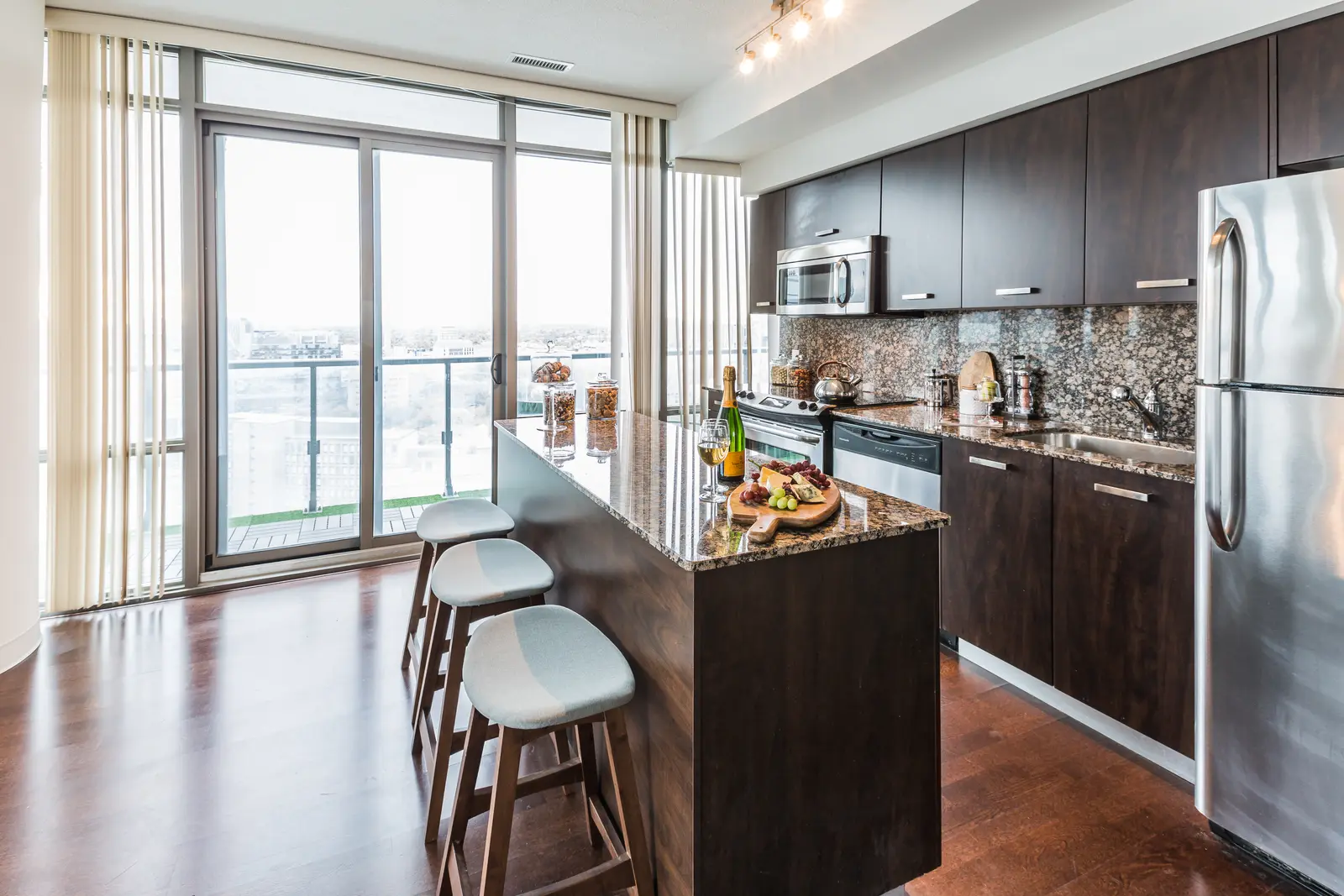 Interior of a Bay Street condo in Toronto, with a grey sofa, a dark kitchen, and city windows.