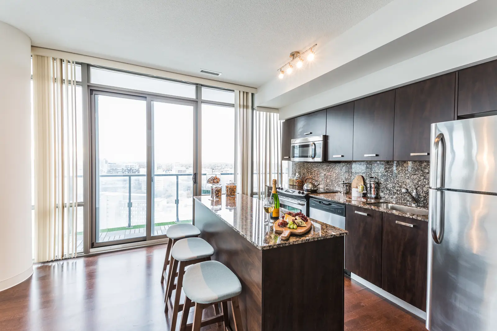 Interior of a Bay Street condo in Toronto, with a grey sofa, a dark kitchen, and city windows.