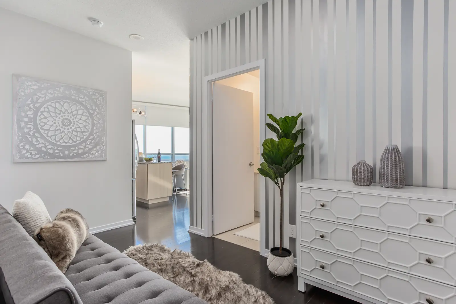 Interior of a waterfront condo in Toronto, with a grey sofa, a light kitchen, and a view of the lake.