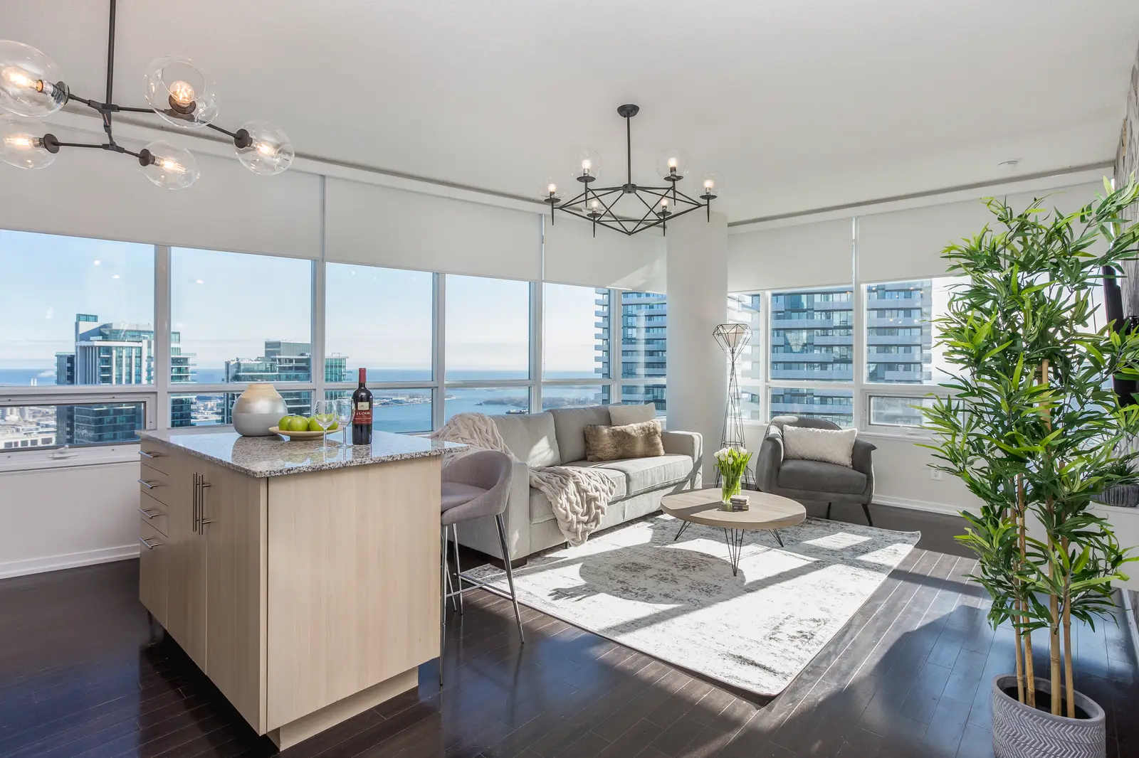 Interior of a waterfront condo in Toronto, with a grey sofa, a light kitchen, and a view of the lake.