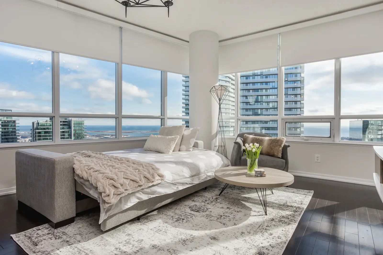 Interior of a waterfront condo in Toronto, with a grey sofa, a light kitchen, and a view of the lake.